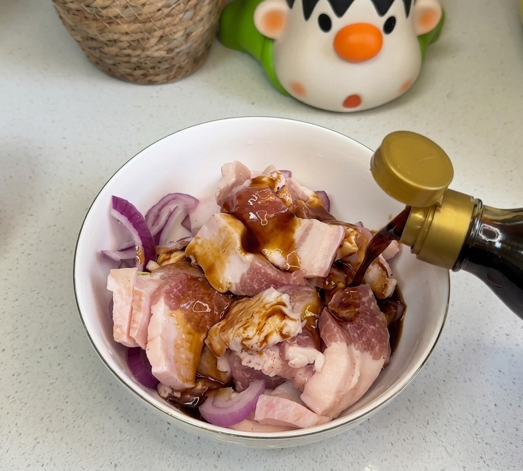 ボウルに入った生の豚バラ肉と赤玉ねぎの上から、醤油やオイスターソースなどの濃い色の液体調味料を注いでいる様子。