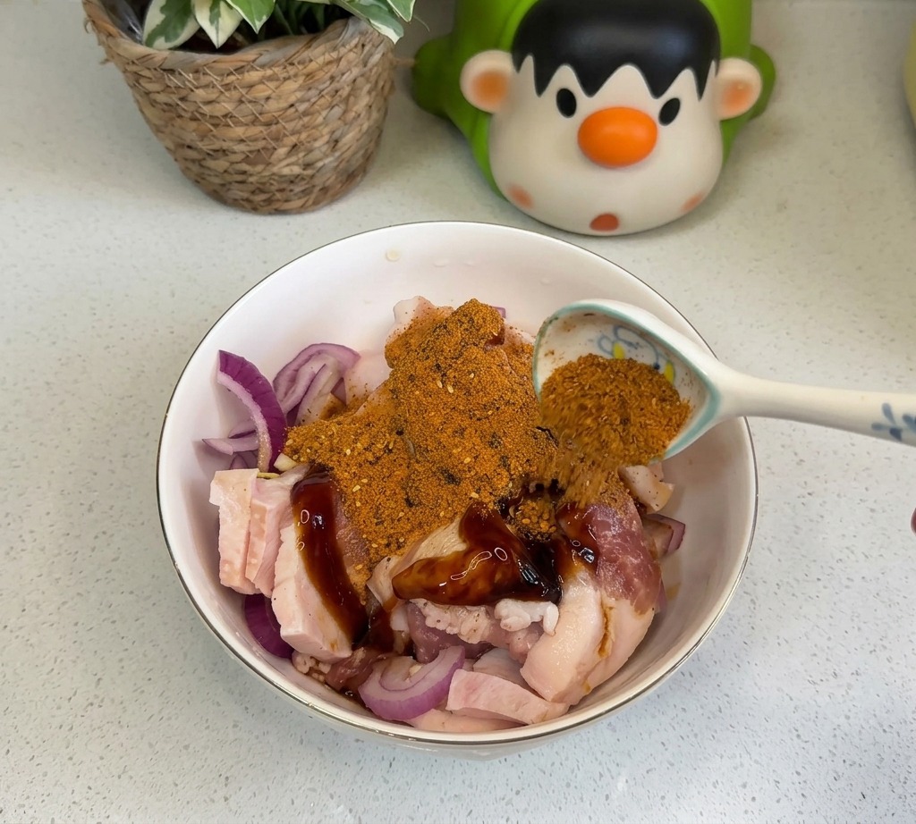 下味をつけた豚バラ肉と玉ねぎの上に、スプーンでオレンジ色のバーベキューシーズニングと赤チリパウダーを振りかけている様子。