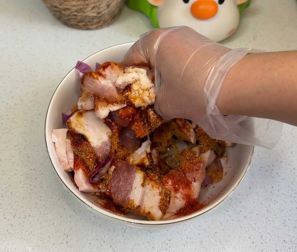 透明な使い捨て手袋をはめた手で、ボウルの中の生の豚バラ肉、玉ねぎ、そして色鮮やかな調味料をしっかりと混ぜ合わせている様子。