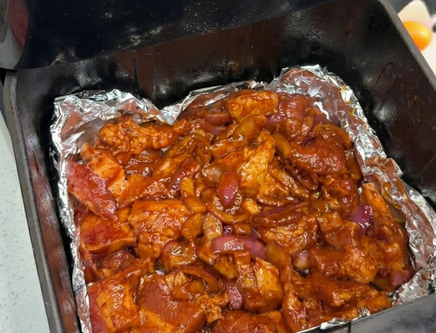 アルミホイルを敷いたノンフライヤーのバスケットの底に、味付けされた豚バラ肉と赤玉ねぎが均一に広げられている様子。