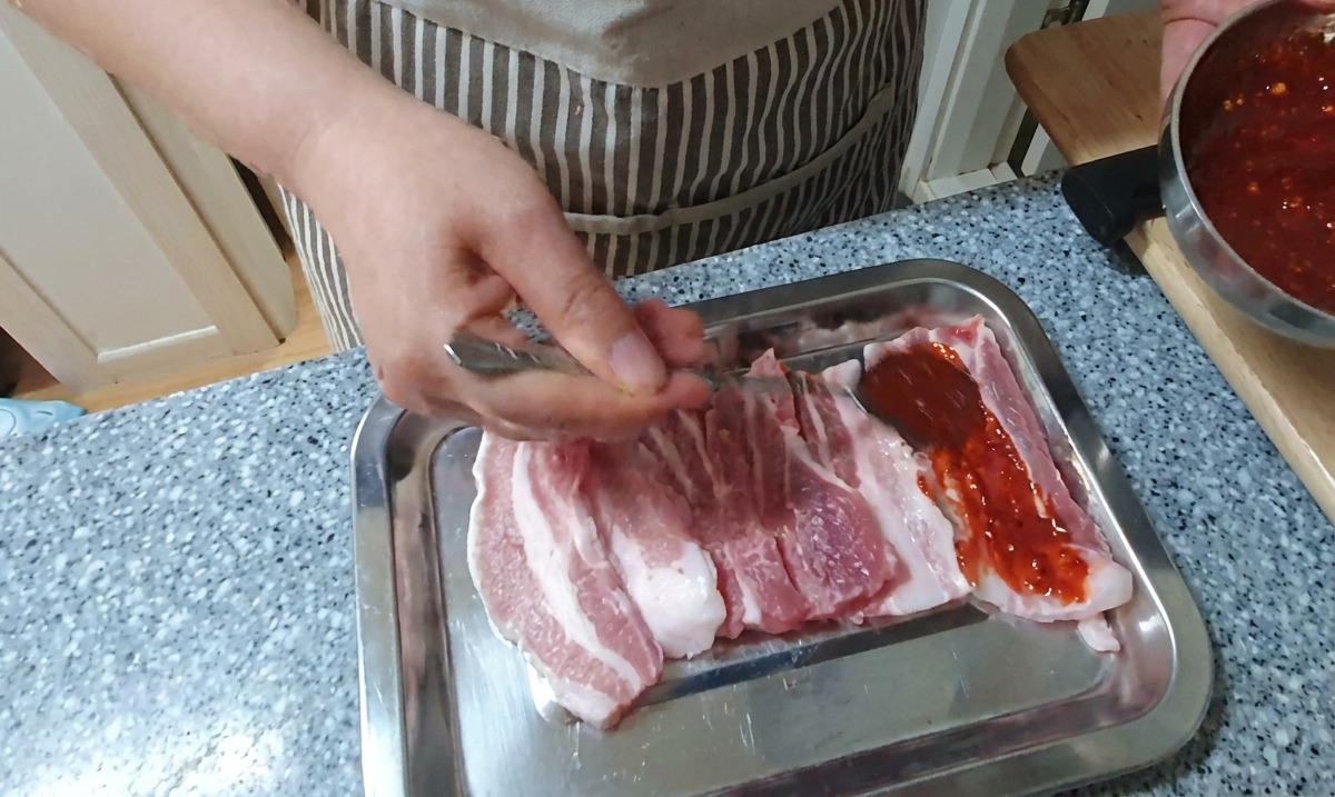 シルバーのトレイの上で、生の豚バラ肉のスライスにスプーンで真っ赤な濃厚コチュジャンだれをまんべんなく塗っている手元の様子