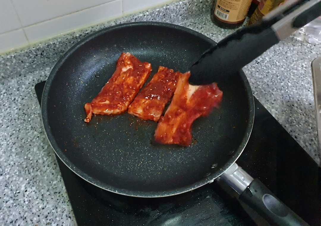 トングを使って、タレに漬け込んだ真っ赤な豚バラ肉を黒いノンスティック加工のフライパンに並べている様子