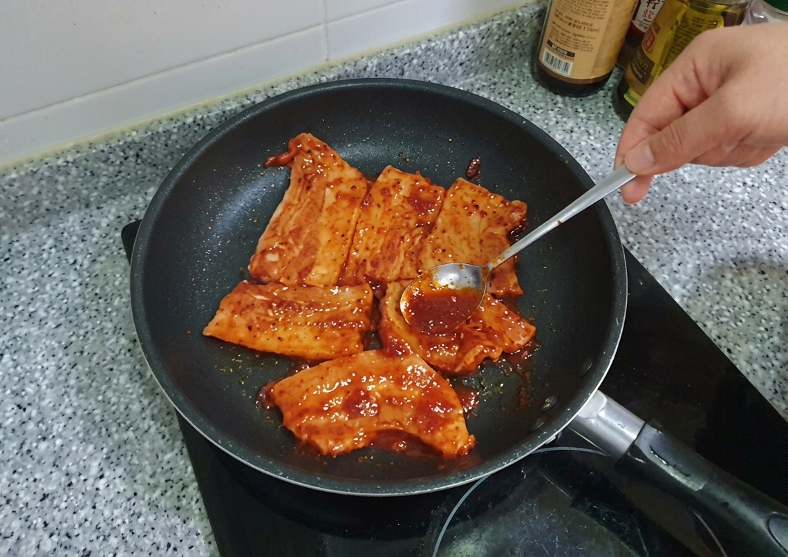 フライパンの中の豚バラ肉の上に、スプーンで濃厚な赤色のコチュジャンだれを塗り広げている様子