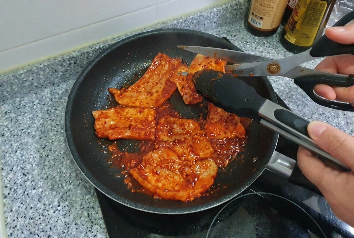 フライパンの中で、キッチンバサミを使ってピリ辛に焼き上がった豚バラ肉を一口大にカットしている様子