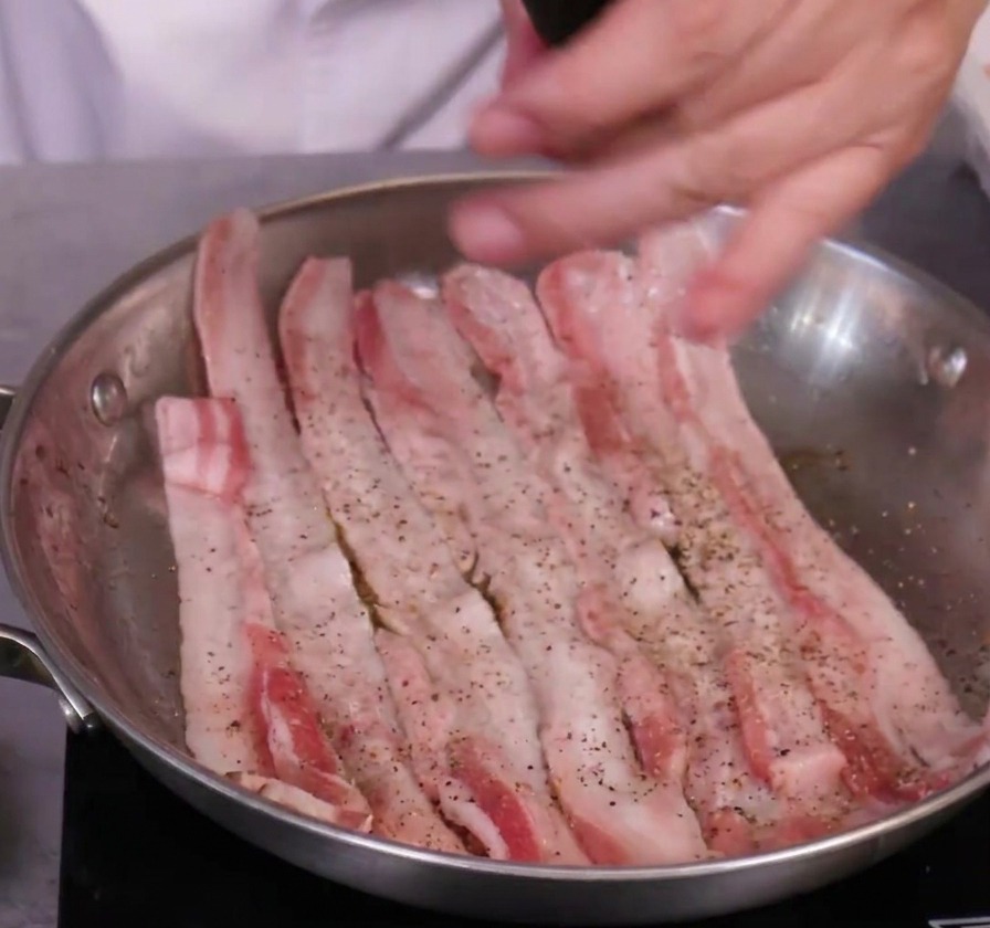 ステンレス製のフライパンの中で、ペッパーミルを使って生の豚バラ肉に黒コショウを振っている様子。
