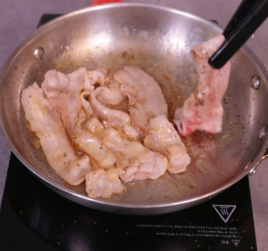 ジューシーに音を立てるフライパンの中で、黒いトングを使ってきつね色に焼けた豚バラ肉をひっくり返している様子。