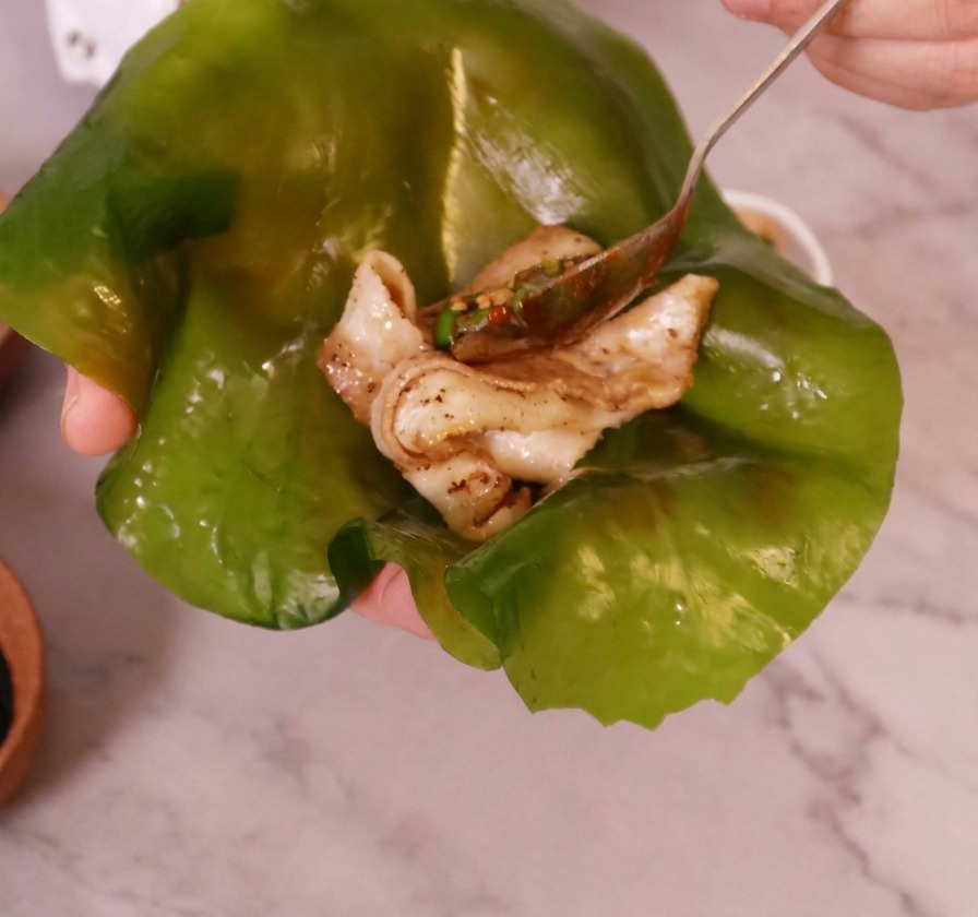 手の上に広げた、やや透明感のある緑色の大きな昆布の葉の真ん中に、焼いた豚バラ肉がのせられている様子。