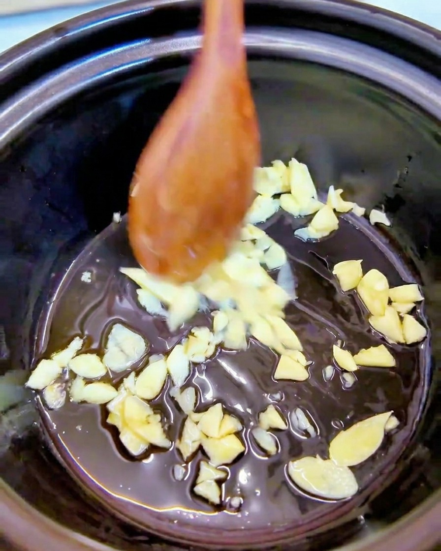 黒い鍋の底で、木のスプーンを使ってごま油とみじん切りのにんにくを炒めている様子。