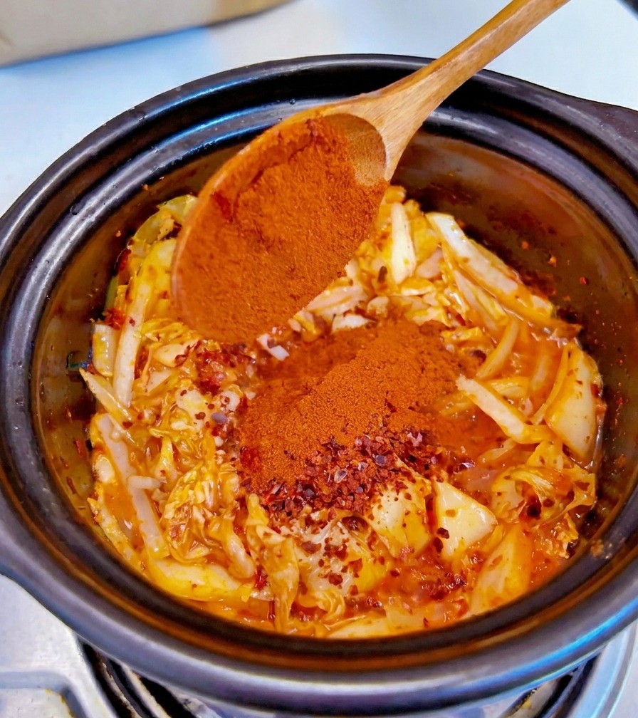 味付けされたキムチと玉ねぎの具材に、木のスプーンから細挽きの赤粉唐辛子が振りかけられている様子。
