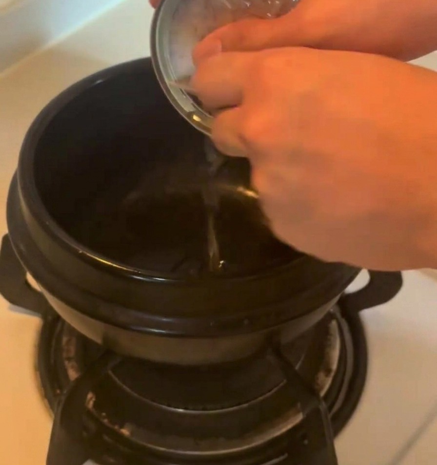 コンロの上の黒い土鍋に、金属製のツナ缶から旨味の詰まったオイルを注いでいる様子。