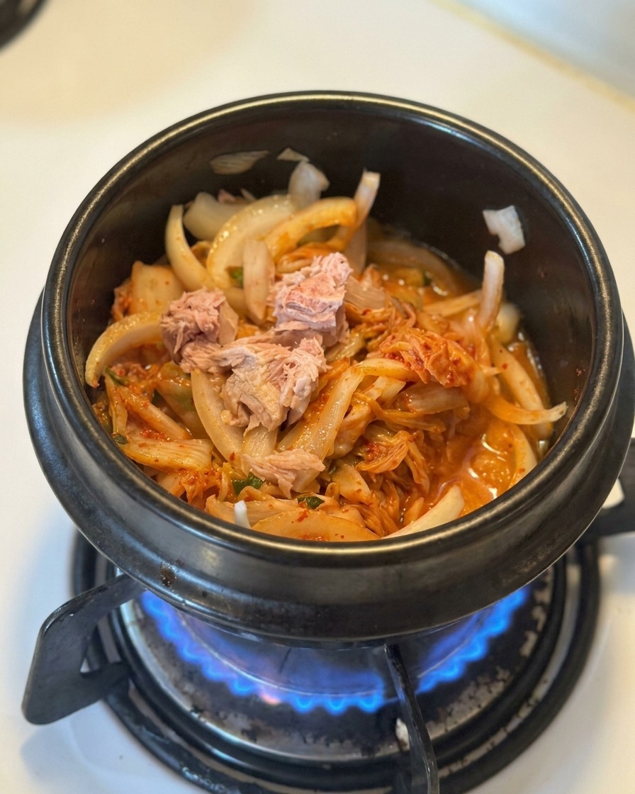 黒い鍋の中で、炒めたキムチと玉ねぎの上に缶詰のツナの塊がのっている様子。