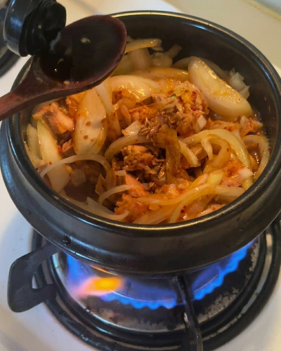 炒めた玉ねぎ、ツナ、キムチが入った黒い土鍋に、木製のスプーンで濃口醤油を注いでいる様子。