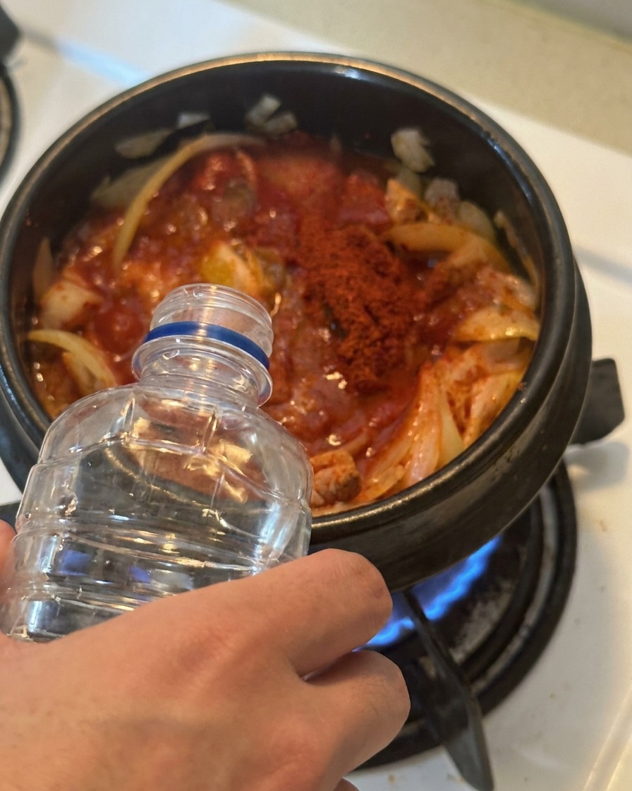 赤く味付けされたキムチとツナが入った鍋に、透明なプラスチックボトルから水を注いでいる様子。
