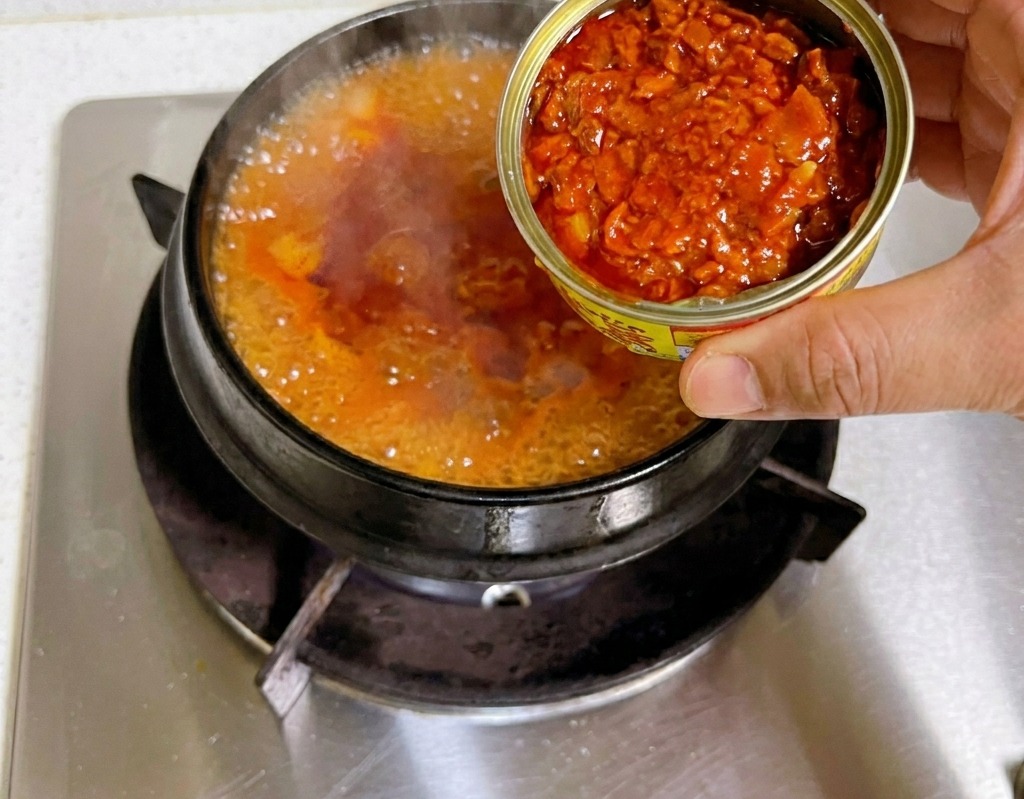 グツグツと沸騰する赤いキムチチゲのベースに、開けたツナ缶からツナをオイルごと入れている様子。