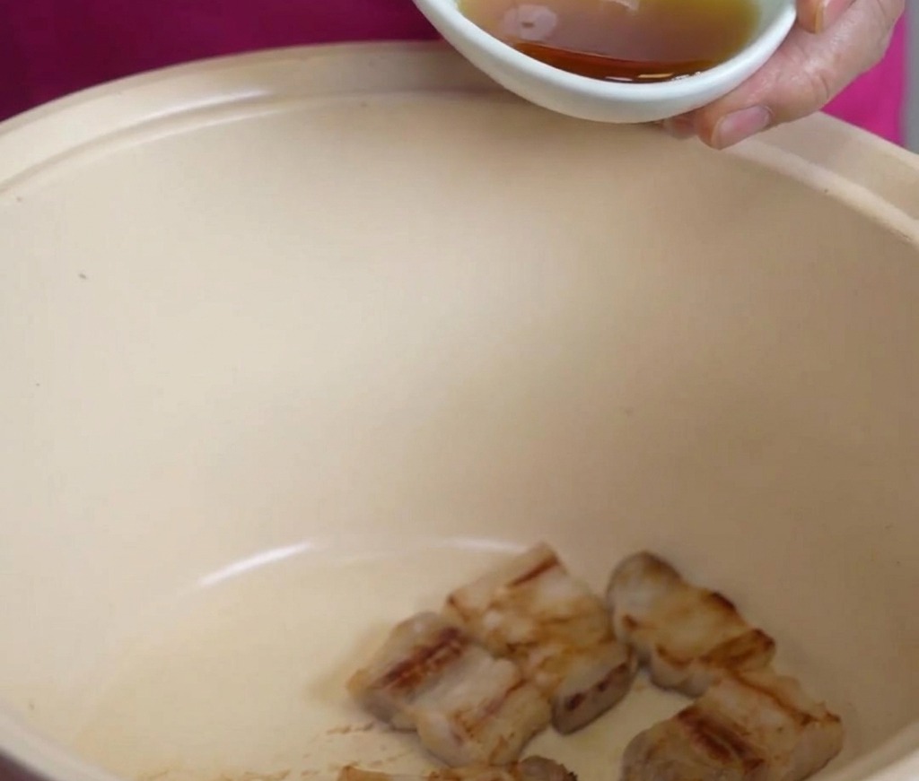 大きめのクリーム色の鍋に入った豚バラ肉の上から、小さな白い器で香ばしいごま油を回しかけている様子