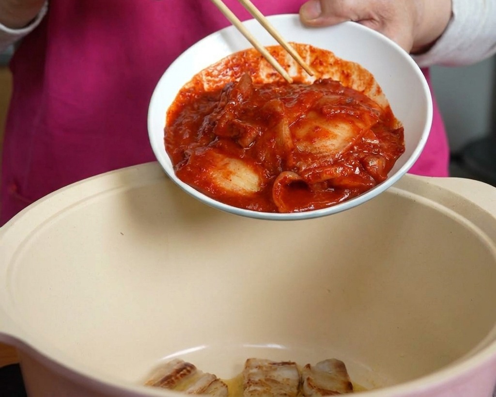 豚バラ肉が入った鍋の中に、箸を使って器から真っ赤で辛そうなキムチを滑り込ませている手元