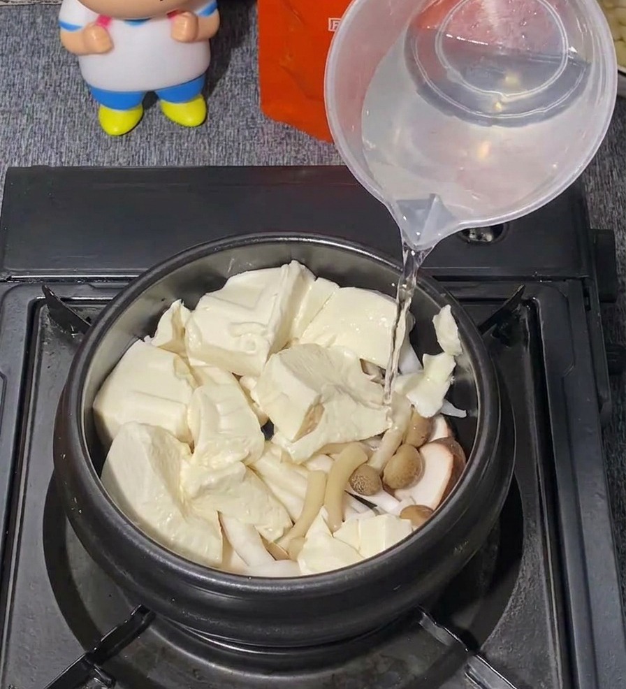 豆腐ときのこが入った黒い鍋に、プラスチックの計量カップからきれいな水を注いでいる様子。