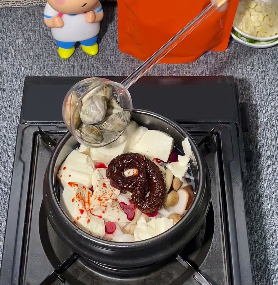 豆腐、きのこ、赤唐辛子の調味料が入った土鍋に、レードルで新鮮なアサリをそっと添えている様子。