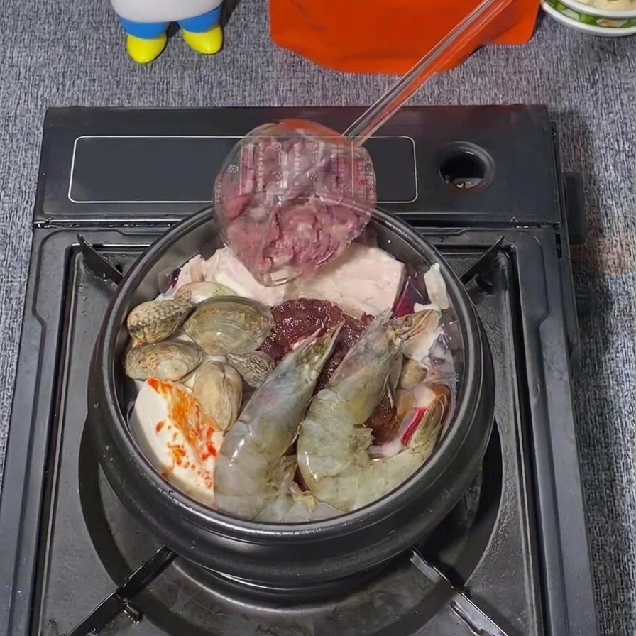 エビやアサリが入った韓国風チゲ鍋の上に、レードルから薄切りの生牛肉をのせている様子。