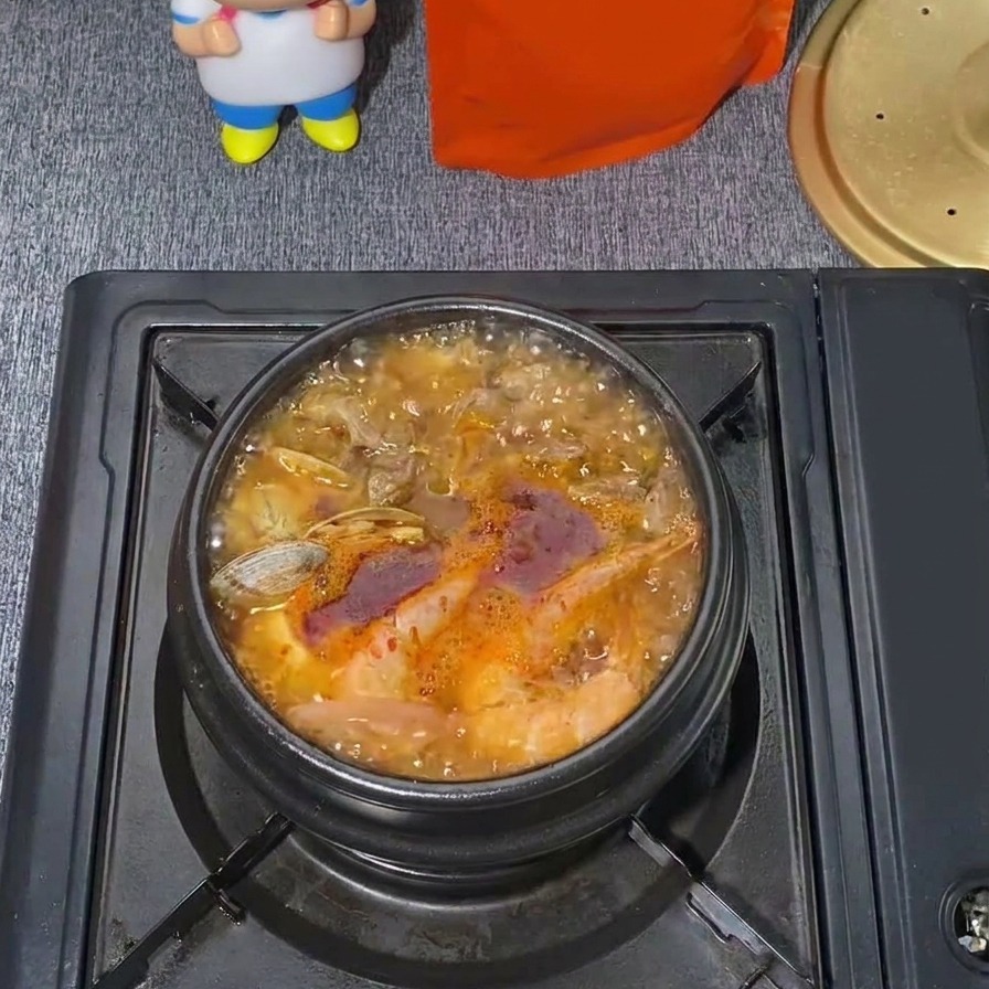 真っ赤なスープが激しく沸騰し、アサリの殻が開き、湯気が立ち上っている韓国風海鮮スンドゥブチゲの鍋。