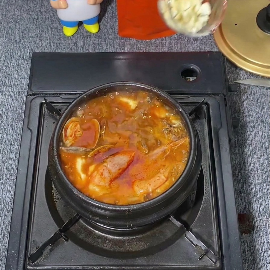 沸騰したピリ辛の赤いチゲの真ん中に、透明なレードルからすりおろしニンニクを1スプーン落としている様子。