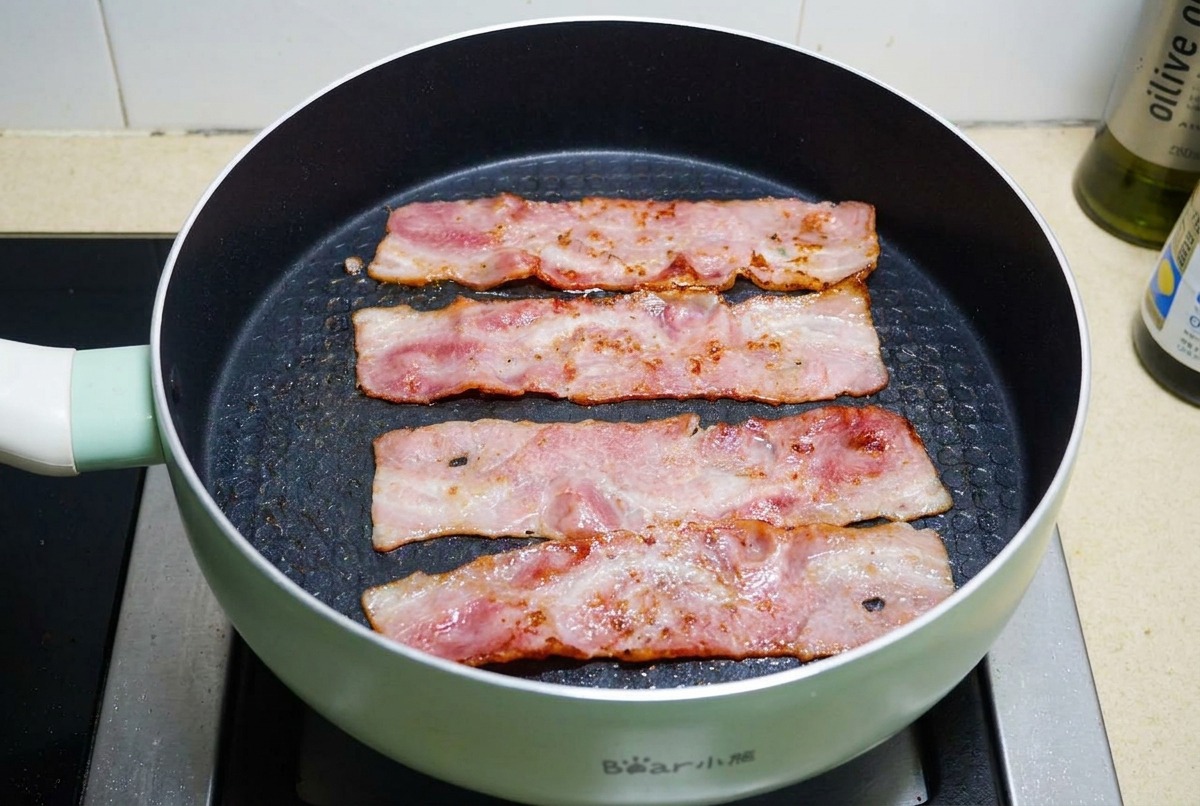 黒いフライパンで、脂が泡立ち表面がこんがりとカリカリになるまで焼かれている4枚のベーコン。