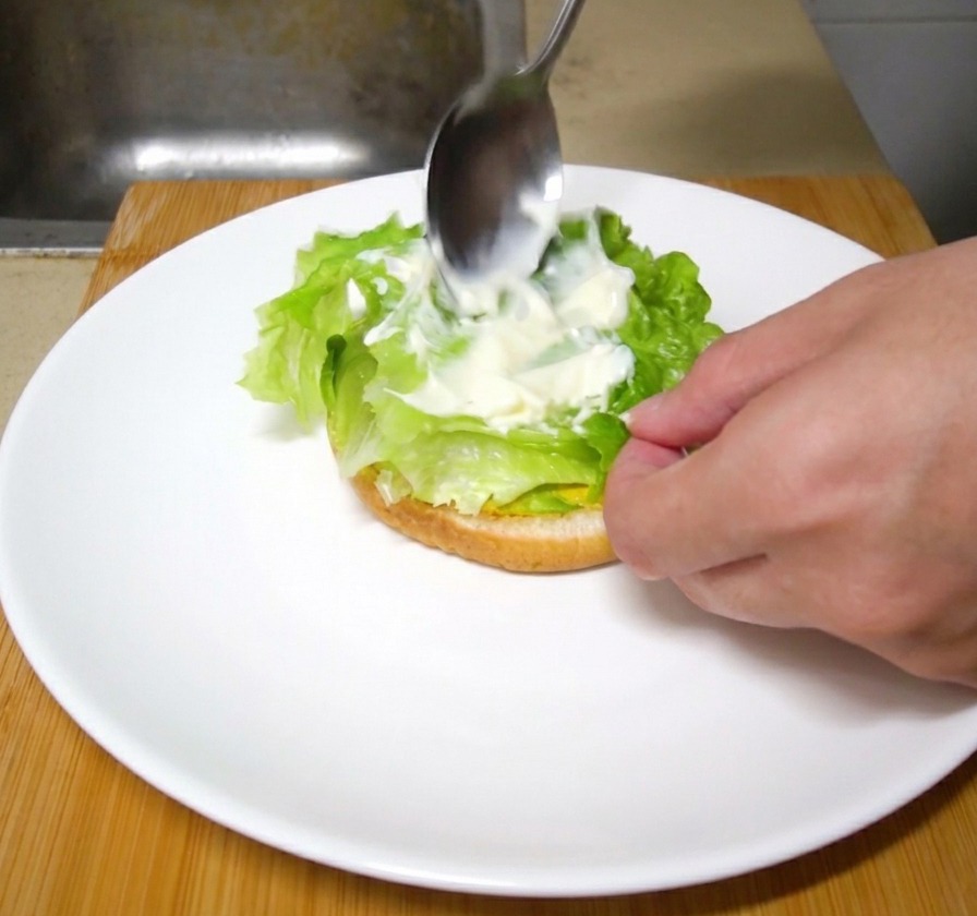 バーガーの層に重ねられた新鮮なレタスの上に、金属製のスプーンで白いマヨネーズを塗り広げている様子。