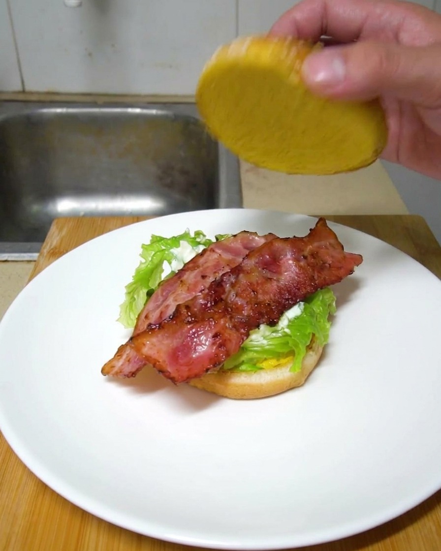 レタスとカリカリのベーコンがのったお皿のバーガーに、上のバンズを手でそっとかぶせている様子。
