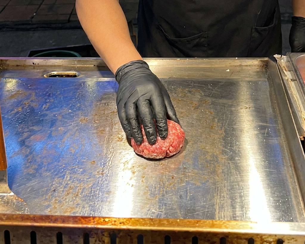 黒い手袋をはめた手が、熱い鉄板の上で丸い生の牛ひき肉を金属製のプレスで平らなパティ状に押し潰している様子。