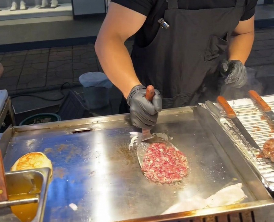 業務用の鉄板の上で、炒めた赤タマネギの山の上に金属製のスパチュラでビーフパティをひっくり返して重ねる様子。