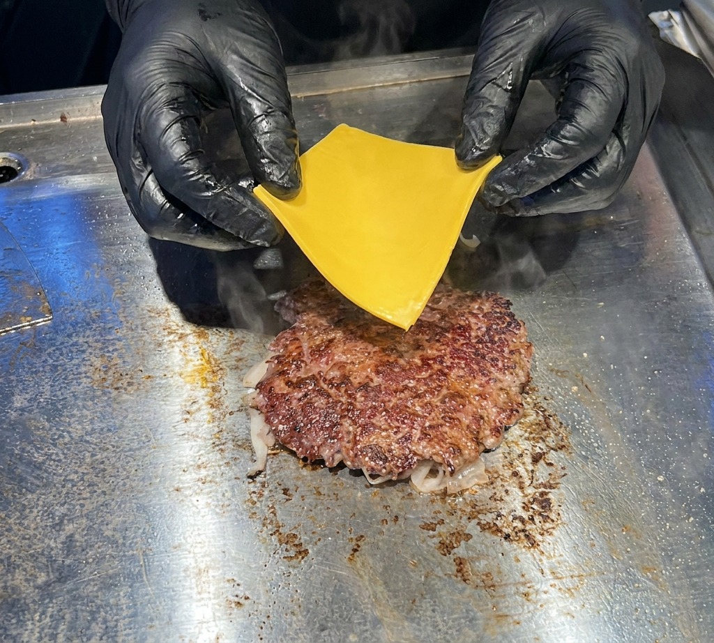 平らな鉄板の上でジュージューと音を立てるこんがり焼けたビーフパティの上に、黒い手袋をはめた手が鮮やかな黄色のチェダーチーズを丁寧にのせている接写。
