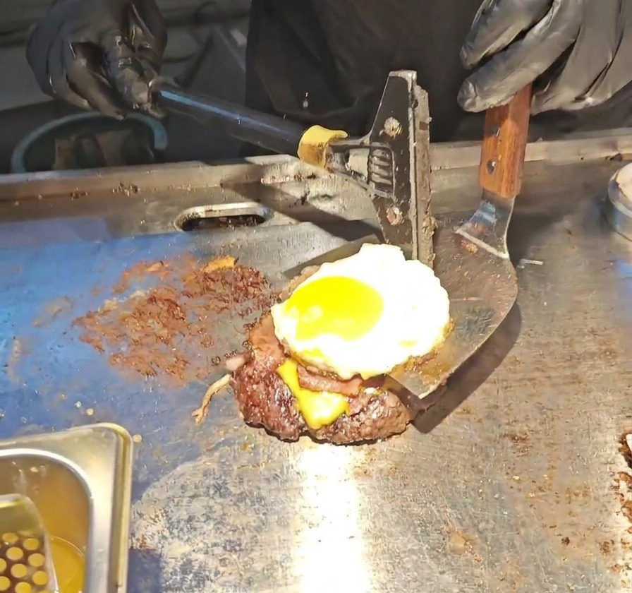 ビーフパティ、とろけたチーズ、ベーコン、そして半熟の目玉焼きが重なったボリュームのあるバーガースタックを、幅広の金属製スパチュラで持ち上げている様子。