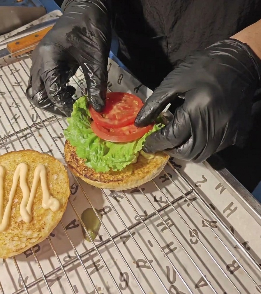 黒い手袋をはめた手が、トーストしたバーガーバンズの上の緑色のレタスの上に、新鮮なトマトのスライスをのせている様子。
