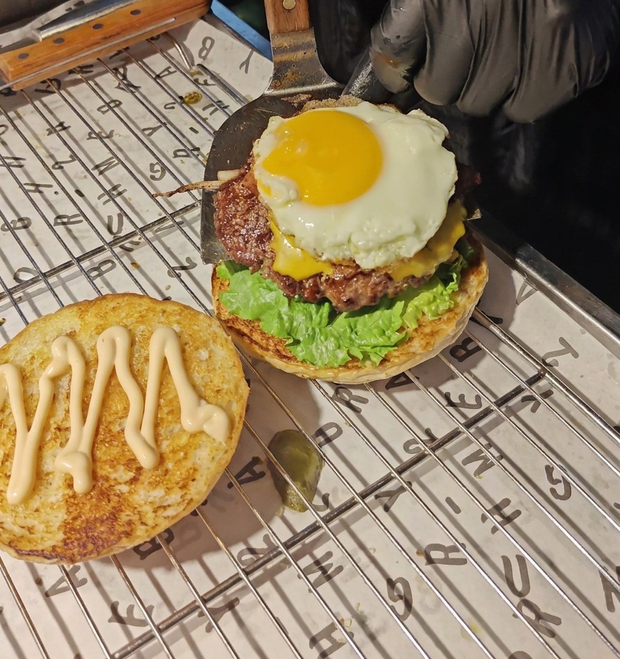 金属製のスパチュラを使って、とろけたチーズと目玉焼きがのったビーフパティを、レタスとトマトの層の上に慎重に移している様子。