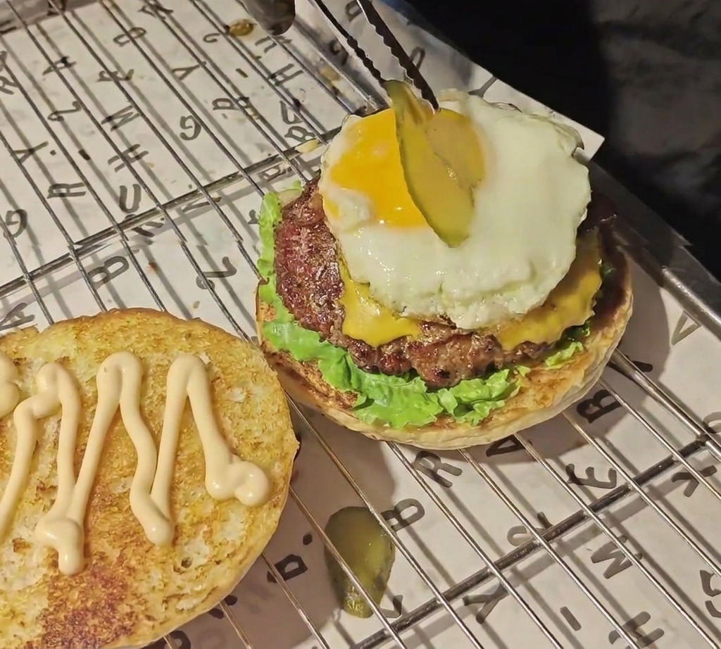 トングを使って、バーガースタックの頂上にある鮮やかな黄色の卵黄の上に、薄切りにしたピクルスをのせている様子。