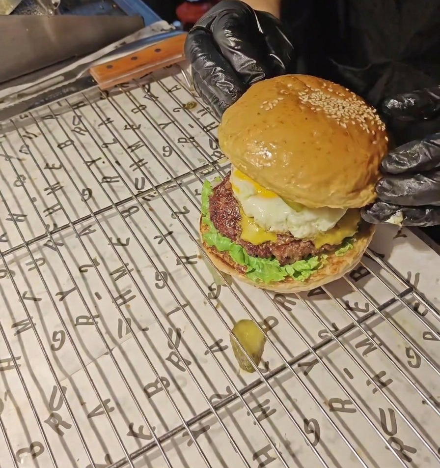 黒い手袋をはめた手が、完成したベーコンエッグバーガーの頂点に、ゴマのついた香ばしいバンズをのせている様子。