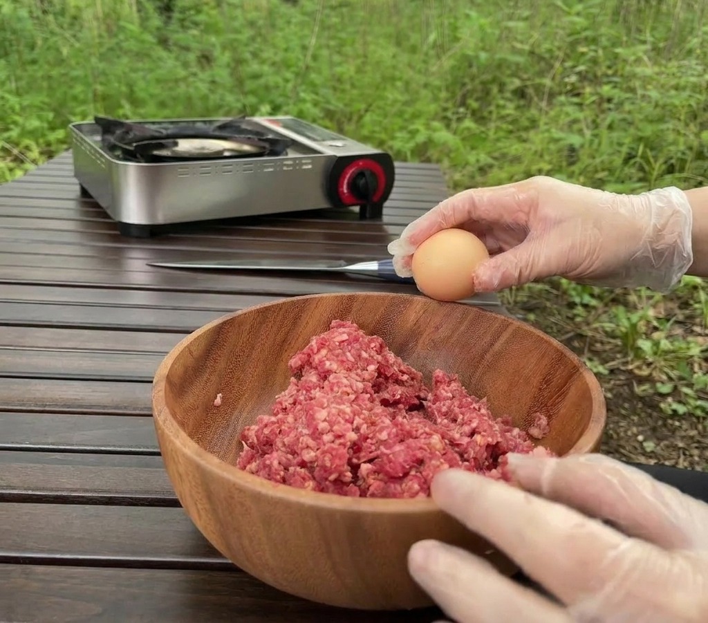 アウトドアの木製テーブルの上で、牛ひき肉が入った大きな木製ボウルに向かって、透明な手袋をはめた手で生卵を割り入れようとしている様子。