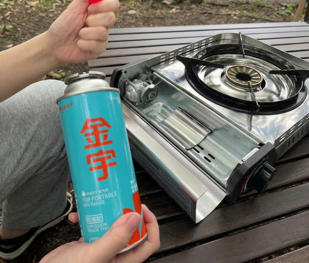 開いたポータブルコンロのガス缶設置スペースの横で、青いカセットガス缶と赤色のキャップを持っている様子。