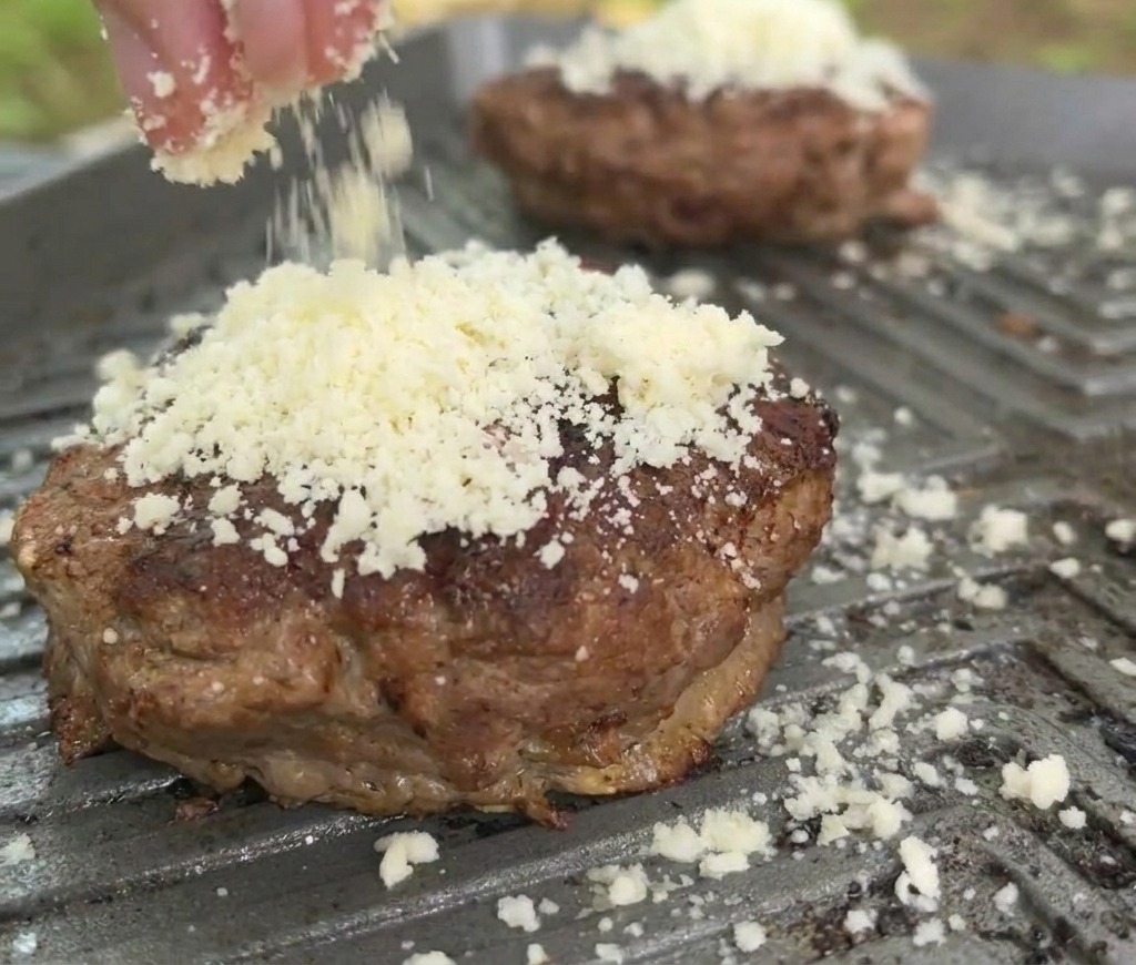 熱いグリルパンの上でこんがりと焼けたビーフパティの上に、たっぷりの白いシュレッドチーズを振りかけているクローズアップ。