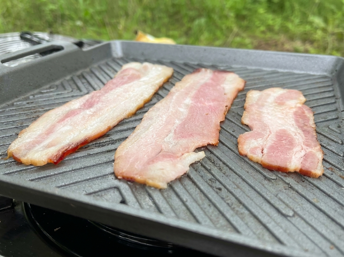 グレーの溝付きグリルパンの上で3枚の長いベーコンが焼かれ、脂がじんわりと溶け出している様子。