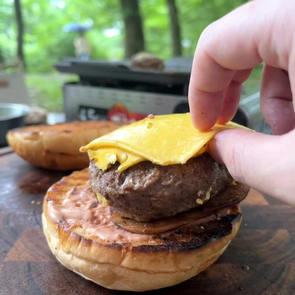 ピンクソースが塗られたトーストバンズの上に、とろける黄色いチーズがのった焼き立てのビーフパティを、手で丁寧に移している様子。