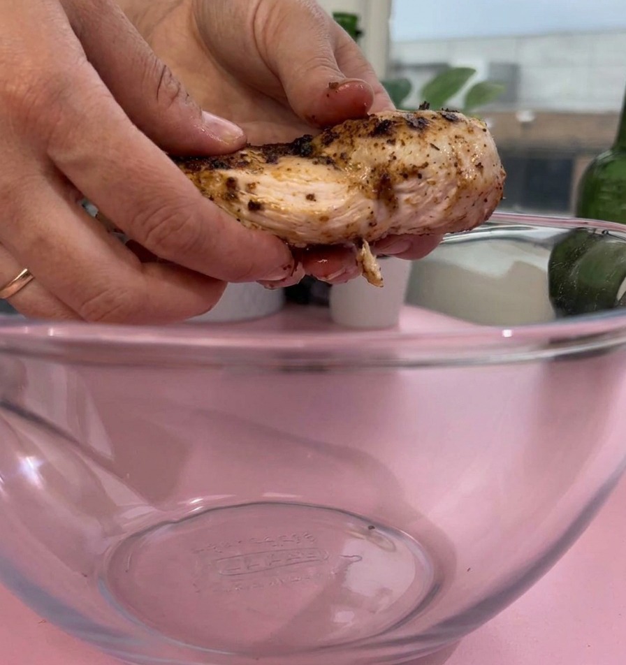 ピンク色の台の上で、味付けして調理された鶏むね肉を透明なガラスボウルに入れている様子。