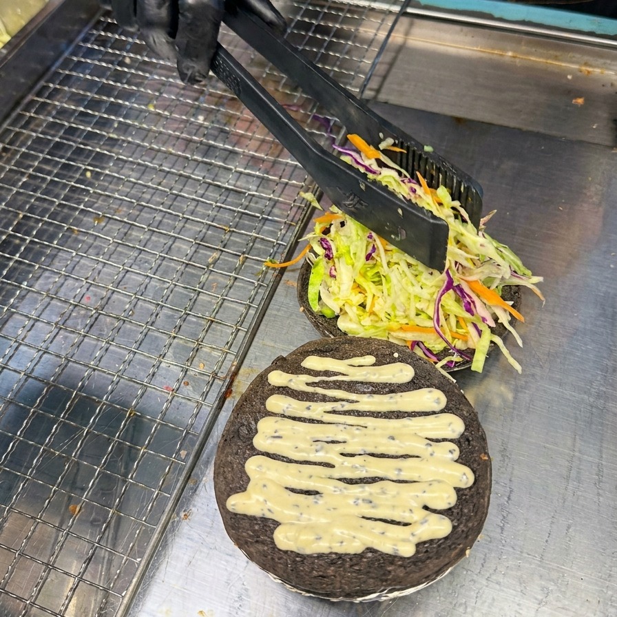 黒いバーズの上の野菜のベッドの上に、トングを使って鮮やかな赤色のトマトスライスを丁寧にのせている様子。