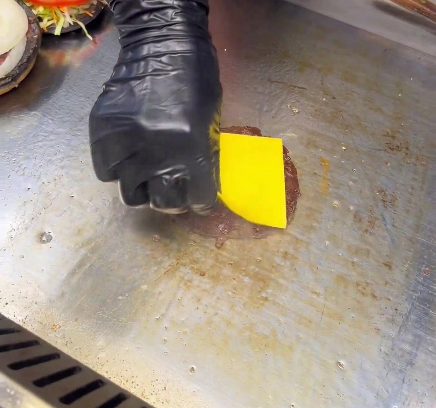 黒い手袋をはめた手が、グリル板の上で焼かれているパティの上に四角いイエローチェダーチーズをのせている様子。