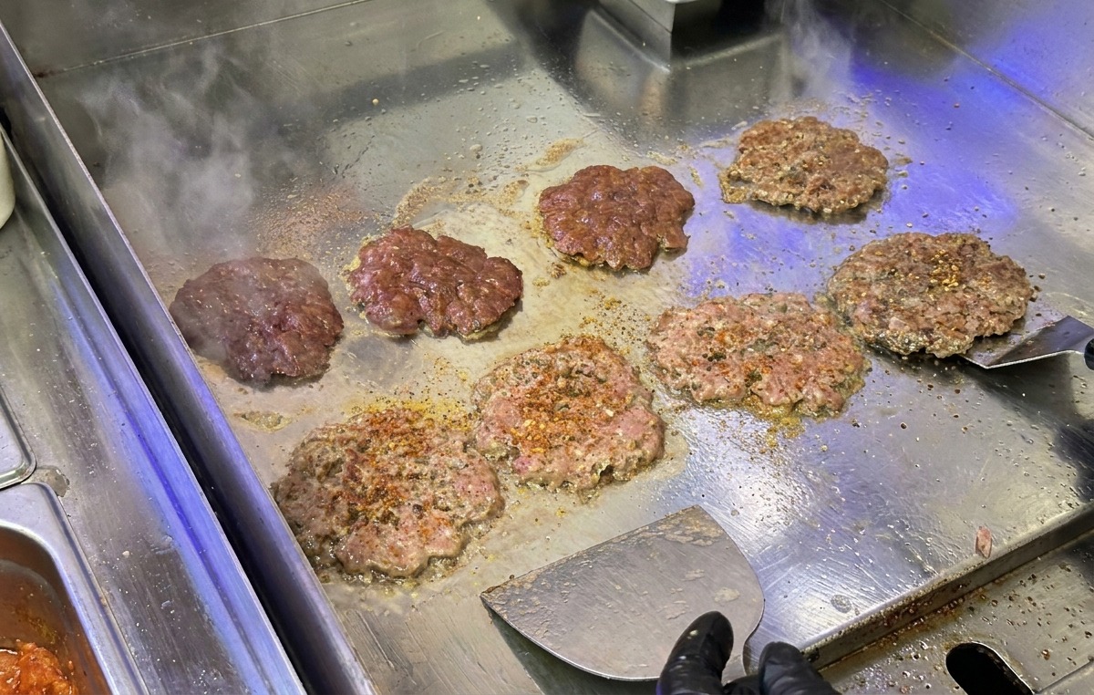 鉄板の上で、金属製のミートプレスやスクレイパーを使って押し付けられながら、香ばしく焼かれているビーフパティ。