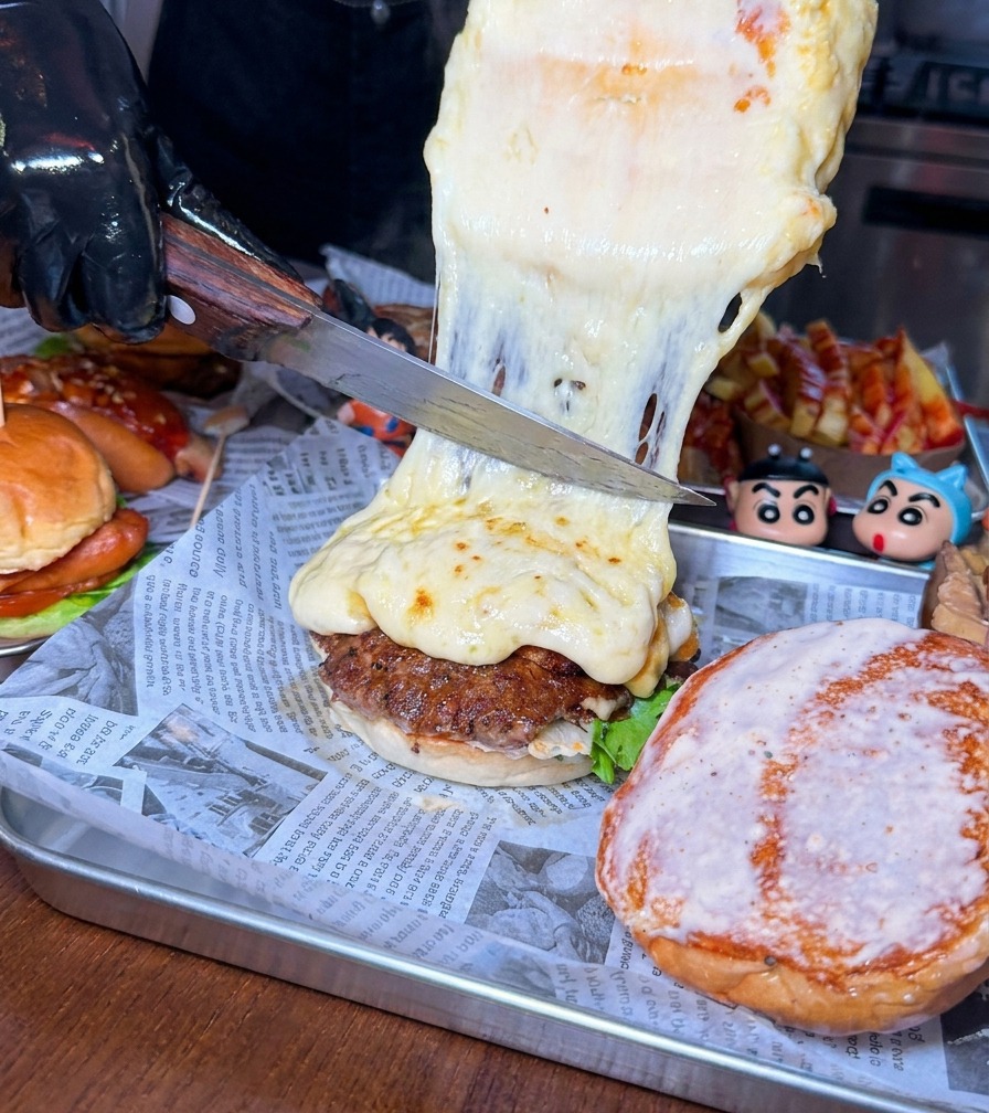 大きなナイフで、ホイールから泡立つ濃厚な溶けチーズをビーフパティに削り落としている仕上げの瞬間。