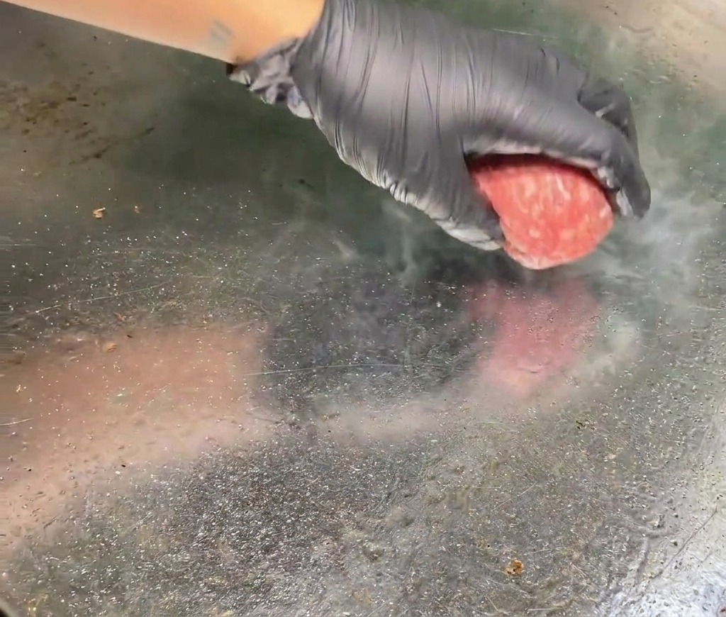 手袋をはめた手が、丸めた生の牛ひき肉を熱い鉄板の上に置き、接触面から湯気が立ち上っている様子。