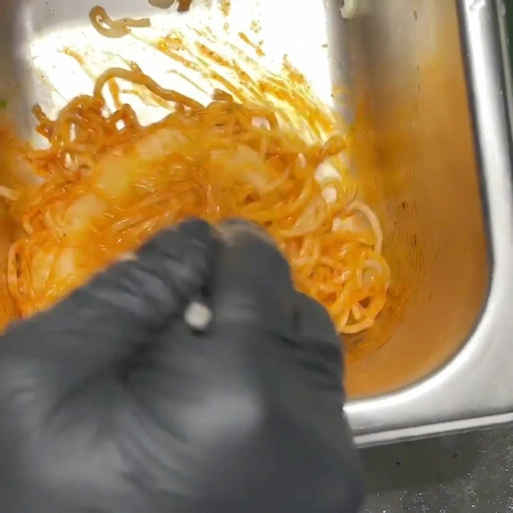 黒い手袋をはめた人が、ステンレス製の容器の中で真っ赤なプルダックソースとラーメンの麺を混ぜ合わせている様子。