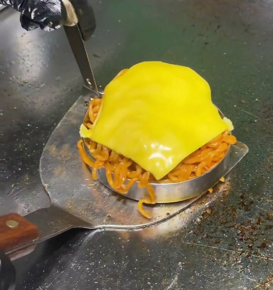プロ用の鉄板の上で、激辛の赤色麺が詰まり、上に黄色いチーズがとろけた丸い型を、金属製のスパチュラで慎重に持ち上げている様子。
