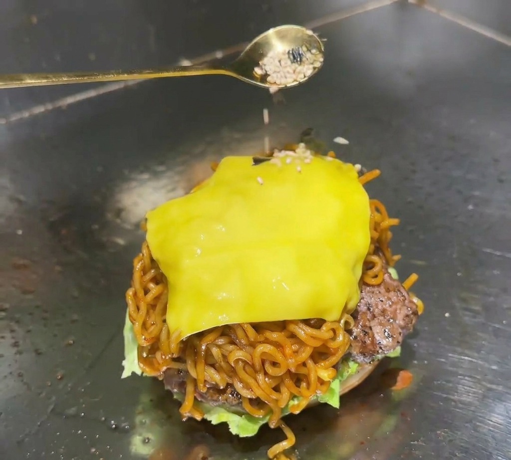 金色のスプーンを使い、ラーメンバーガーのとろけたチーズの上に、白炒りごまと黒い刻みのりのミックスを振りかけている様子。